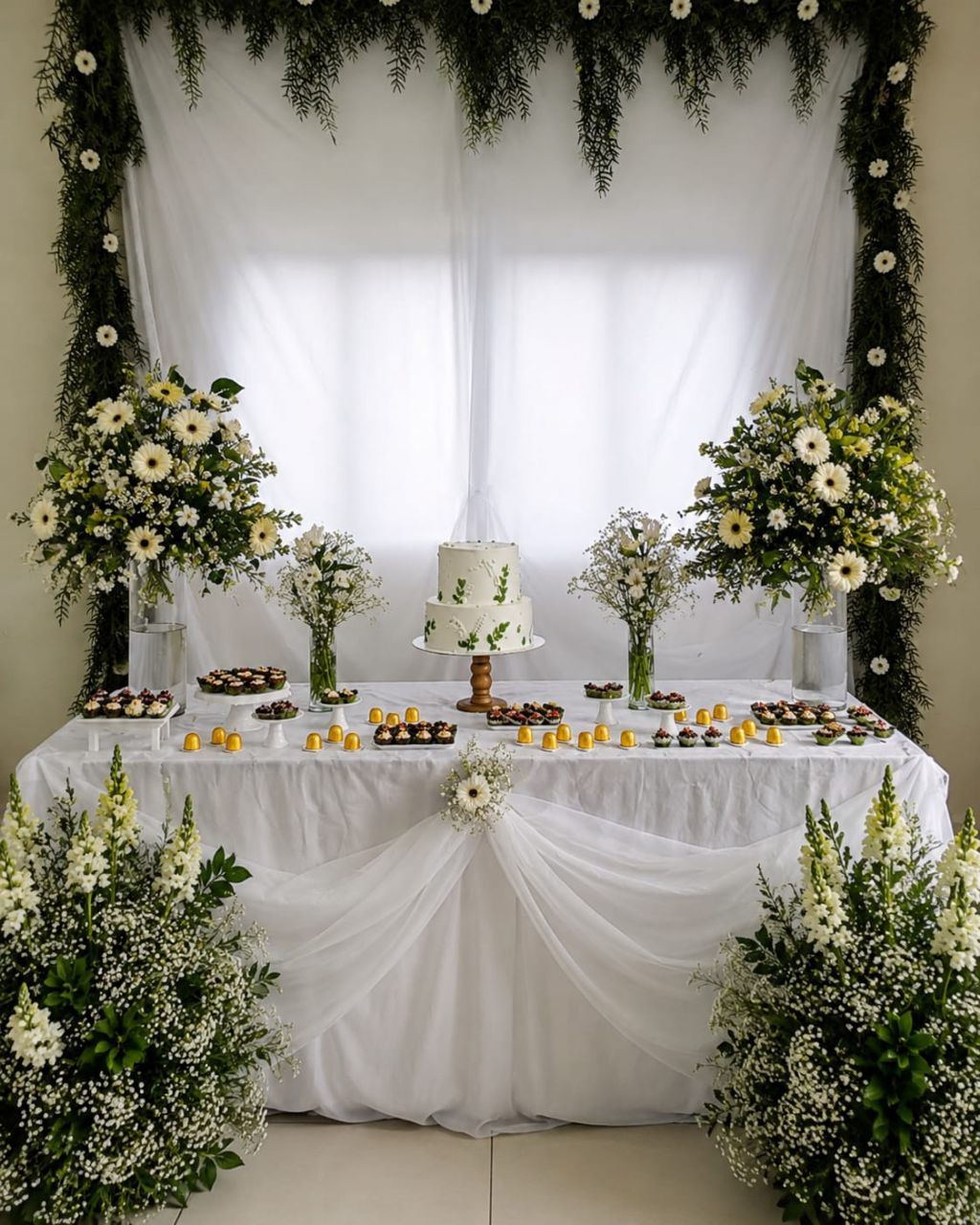 Mesa do bolo decorada com bolo de andares, doces, flores brancas e drapeado em tule
